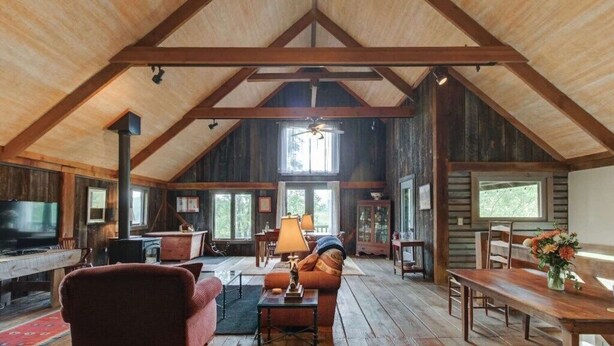 A spacious loft-style room with high ceilings, exposed wooden beams, a large window, a fireplace, and a television.