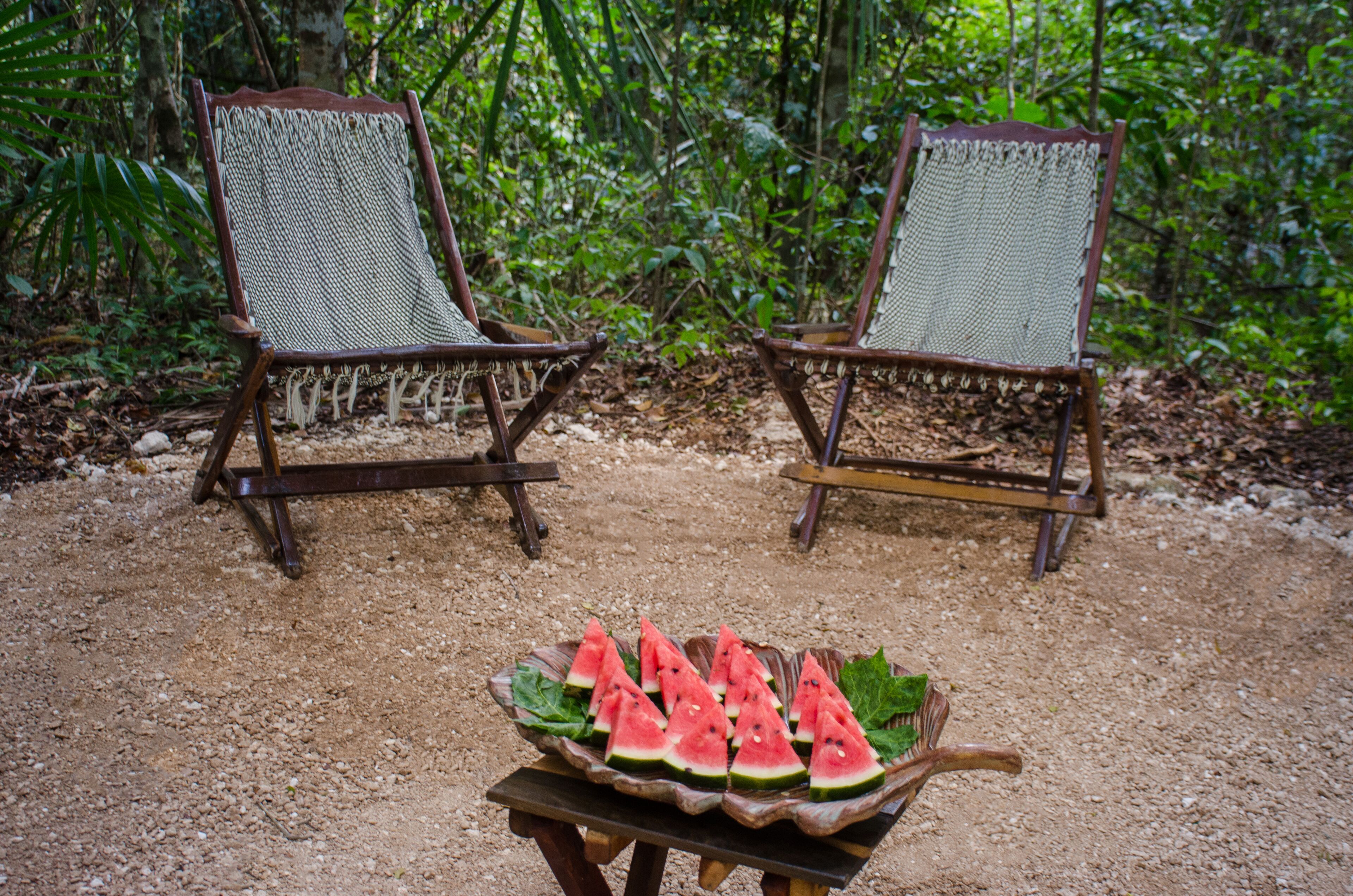 Two wooden chairs with woven seats and backs, placed on a gravel surface with a wooden table in front holding a watermelon dish.