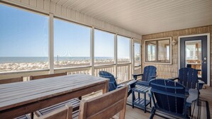 A covered outdoor seating area with wooden tables and chairs, large windows overlooking a beach, and a door leading to an interior space. - A Bella Vista: Coastal Cool Oceanfront Home with Ramp (Oak Island)