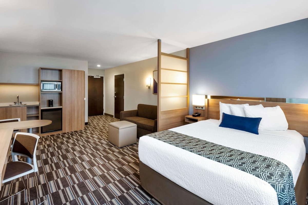 A hotel room with a bed, a desk, a chair, a sofa, a microwave, a coffee maker, and a nightstand with a lamp.