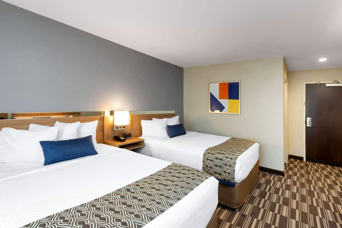A hotel room with two beds, a nightstand with a lamp, a wall art piece, and a door.