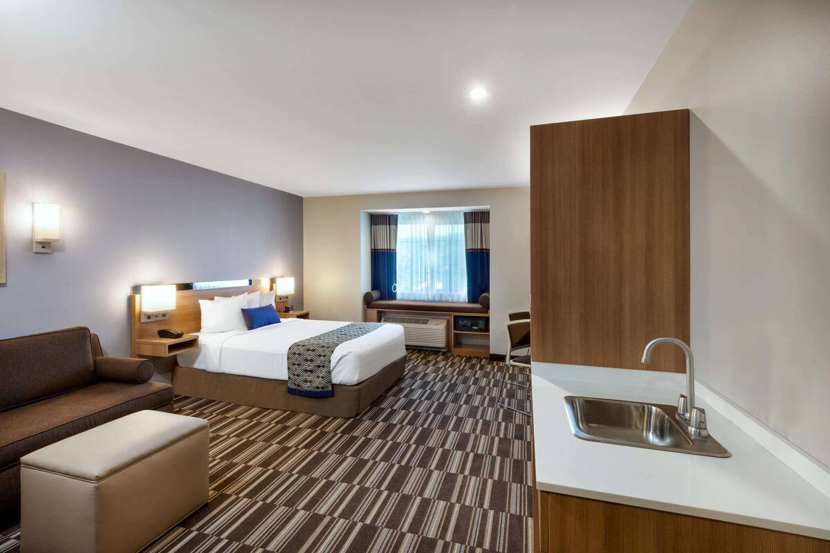 A hotel room with a bed, a sofa, a small table, a desk, a sink, and a window with curtains.