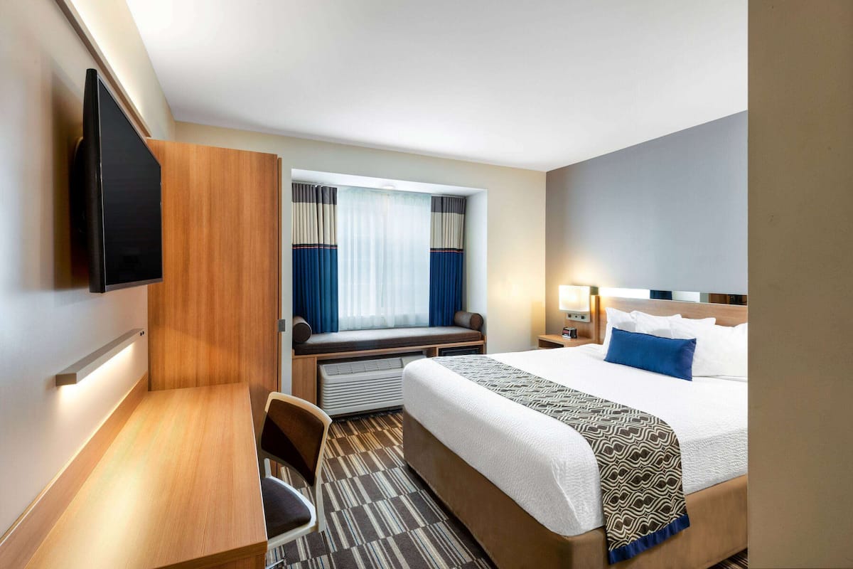 A hotel room with a bed, a desk, a chair, a TV mounted on the wall, and a window with curtains.