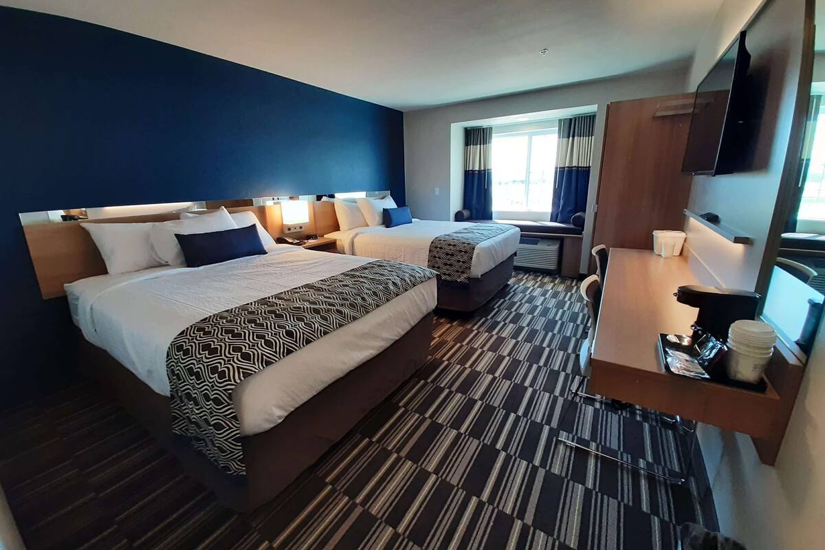 A hotel room with two beds, a desk, a chair, a small table with a cup, and a window with curtains.