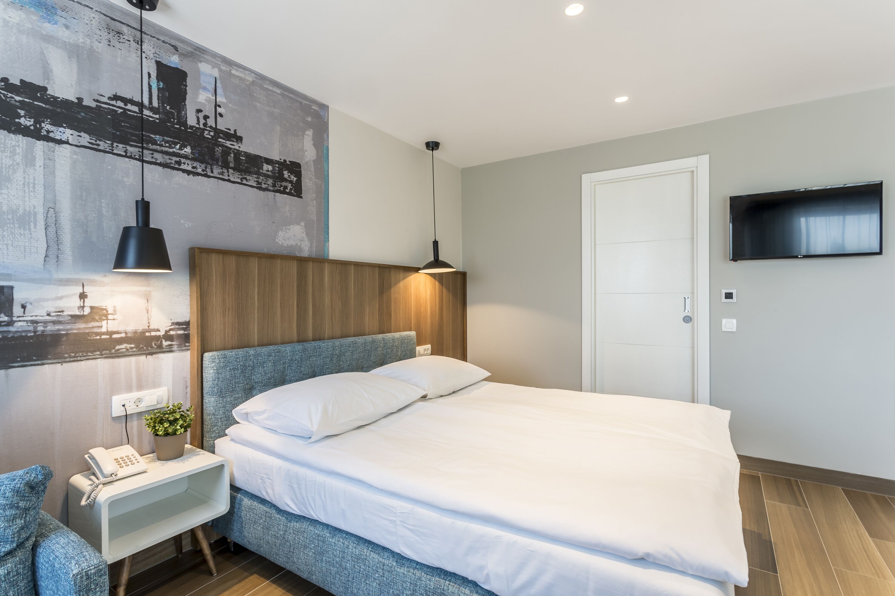 Een moderne hotelkamer met een groot bed, een nachtkastje met telefoon en een plant, een tv aan de muur en een stadslandschap schilderij.