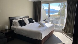 A hotel room with a large bed, a nightstand, a lamp, and a view of a balcony with a table and chairs.