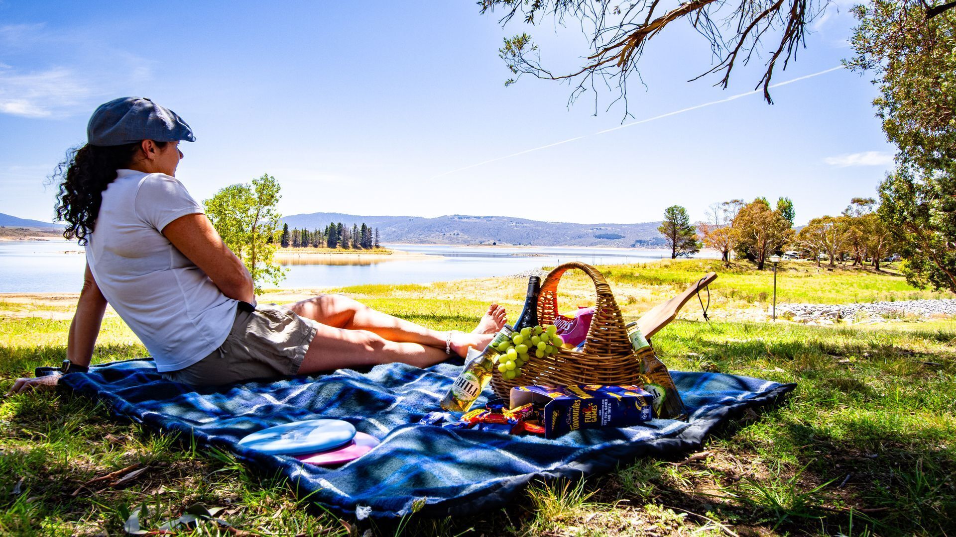 Una persona sentada sobre una manta junto a un lago, con una cesta de picnic que contiene comida y bebidas.