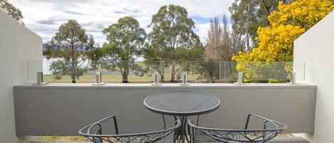 A balcony with a round table and two chairs, overlooking a park with trees and a yellow flowering tree.