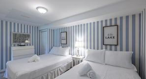 A hotel room with two beds, a bedside table with a lamp, and a wall with striped wallpaper and framed pictures.