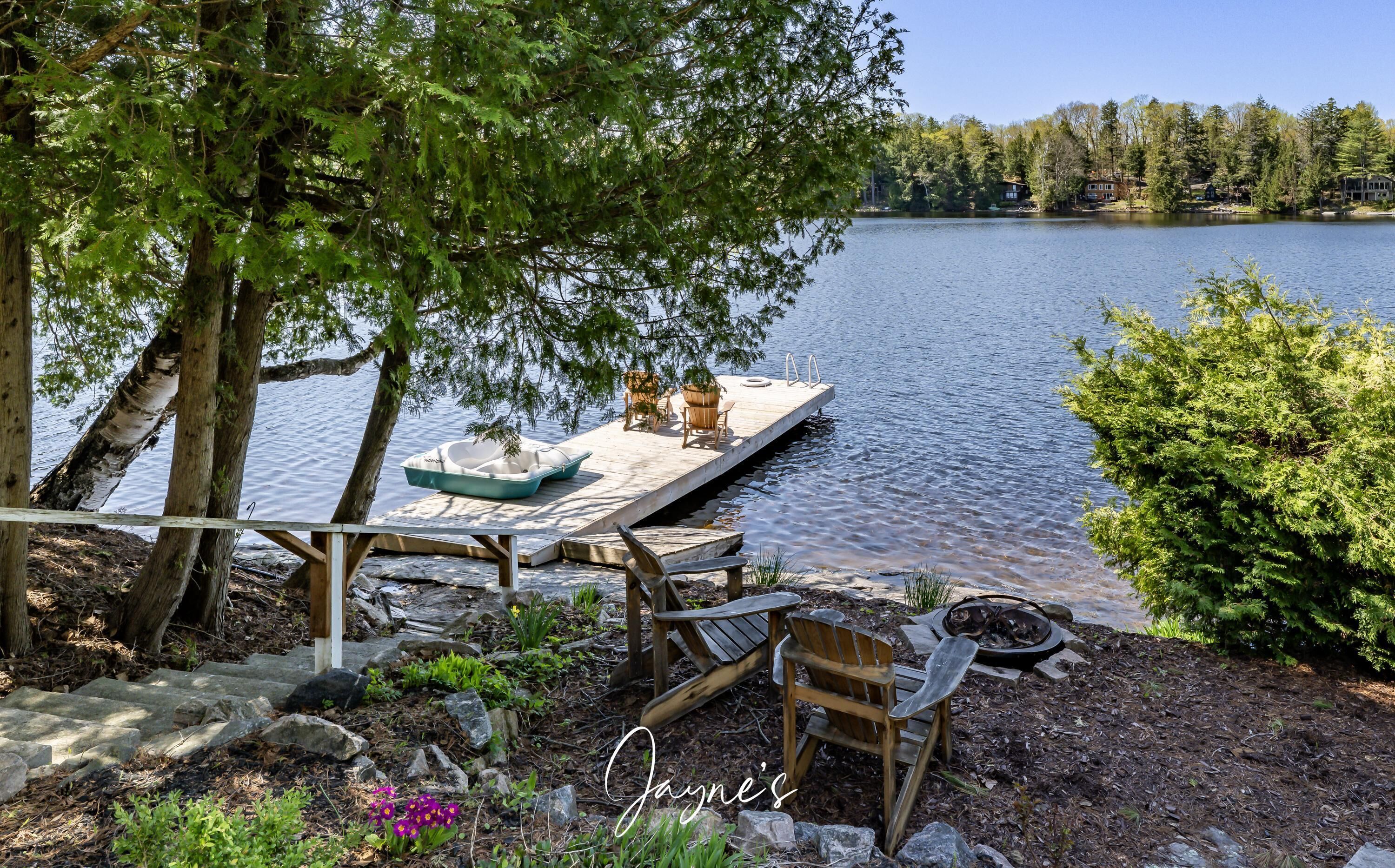 A lakeside dock with a boat, wooden chairs, and a fire pit.