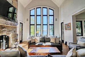 Relax in this spacious living room with a high ceiling and large window offering a view of the outdoors. Features a stone fireplace, comfortable seating area, and a coffee table.