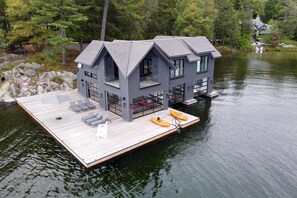 A modern cabin with a wooden deck and kayaks, surrounded by trees and a lake. - AAA Lake Joe Prime Location Riverdale Rd! South! 8bdrm, 5 bath! Muskoka Beauty!! (Port Carling)