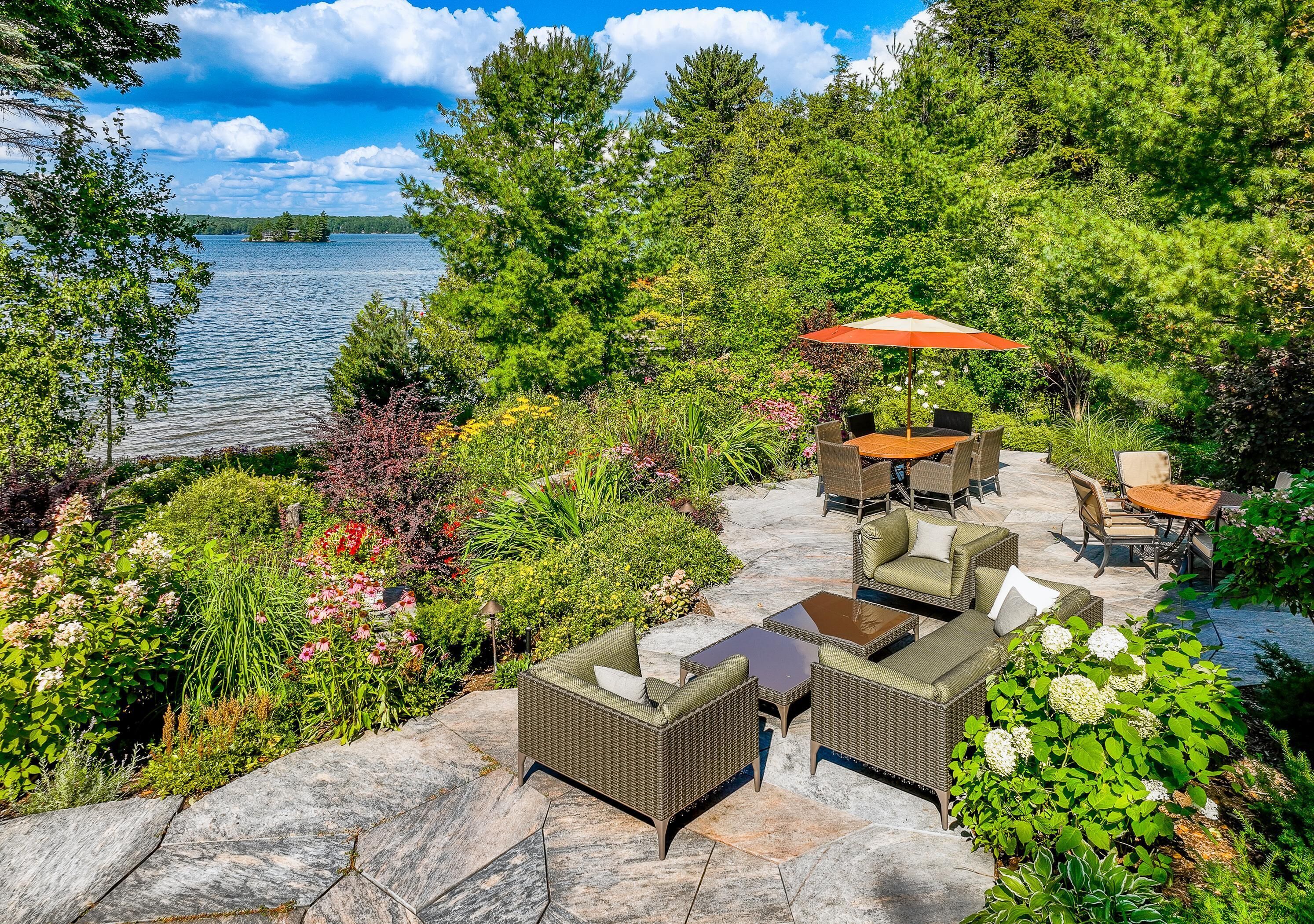 An outdoor patio with wicker furniture, a table, and an umbrella, surrounded by lush greenery and a lake view.