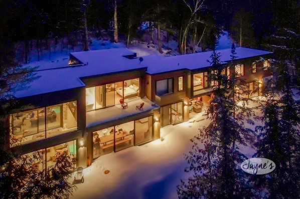 A modern, multi-story house with large windows and a well-lit interior, surrounded by snow.