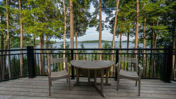 Um deck de madeira com uma mesa e duas cadeiras, com vista para um lago rodeado por árvores altas.
