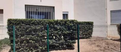 A fenced area with a hedge and a building with barred windows in the background.