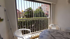 Een balkon met een metalen hekwerk, twee witte plastic stoelen, een ronde tafel met een verenmotief en een witte muur met een klein plankje.
