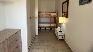 Een smalle gang met een houten stapelbed, een nachtkastje met een lamp, en een spiegel aan de muur.
