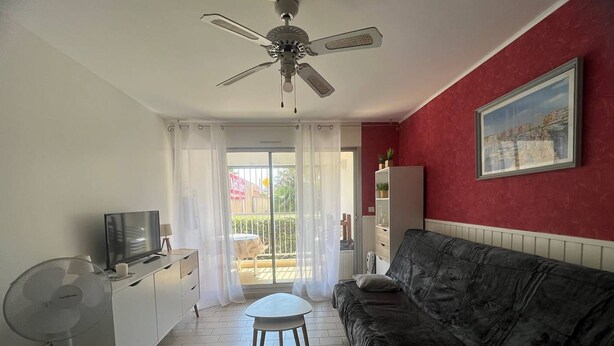 Een woonkamer met een bank, een televisie, een ventilator en een plafondventilator. Er is een schuifdeur naar een buitenruimte.