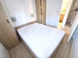 Quarto pequeno com cama de solteiro, cabeceira de madeira, colchão branco e piso de azulejos.