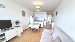 Ein Wohnzimmer mit Fernseher, Ventilator, einer Couch, einem Esstisch mit Stühlen und einer Küche im Hintergrund.