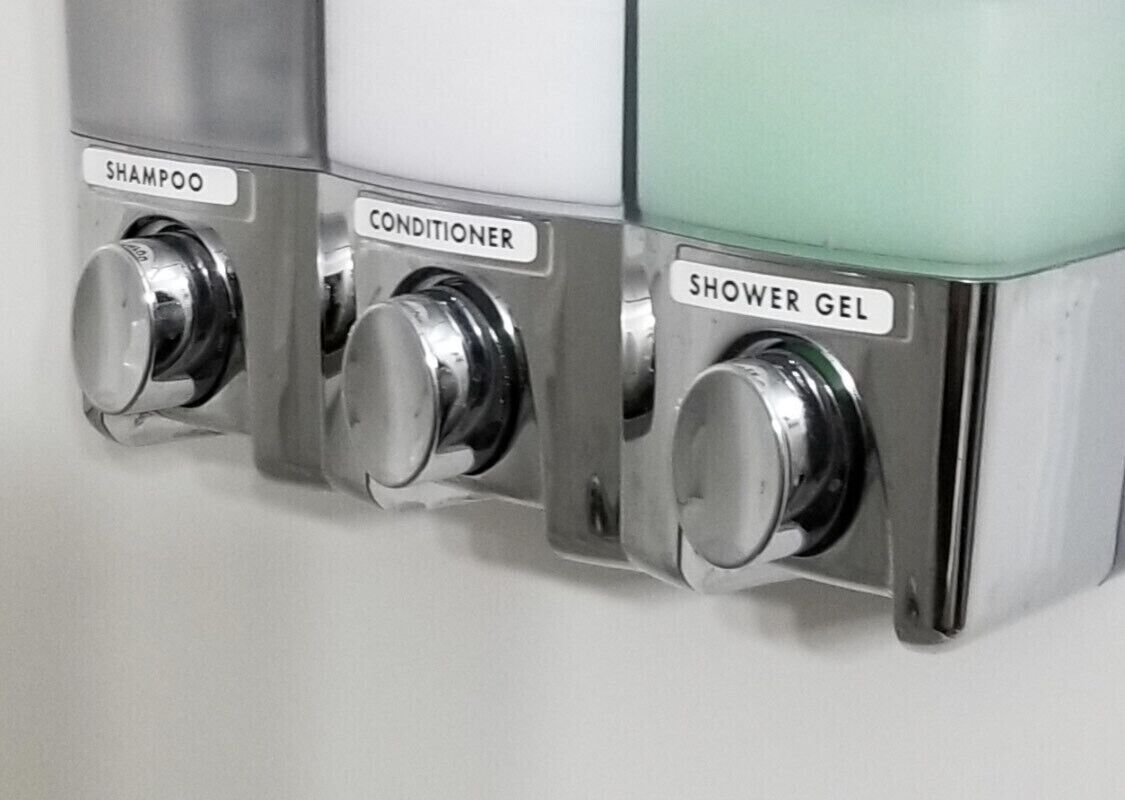 Dispensador montado en la pared con tres compartimentos etiquetados como 'champú', 'acondicionador' y 'gel de ducha'.