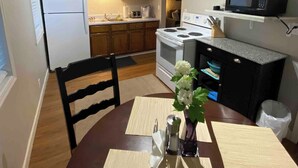 A compact kitchen with wooden cabinets, a white refrigerator, a microwave, and a stove. There is a dining table with a placemat and a vase of flowers. - Skots Villa Bottom East. Centrally located near Sanford Medical Center & Midco. (Sioux Falls)