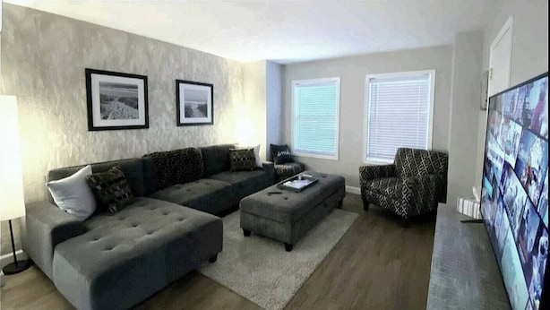 A living room with a grey sectional sofa, an ottoman, a patterned armchair, and a flat-screen TV. - Skots Villa Bottom East. Centrally located near Sanford Medical Center & Midco. (Sioux Falls)