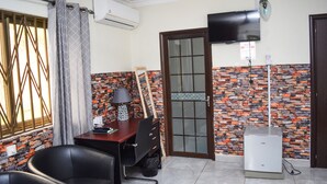 A compact room with a tiled floor, a desk with a lamp, a television mounted on the wall, and a door leading to another room. - 3A's Guest House (Akosombo)