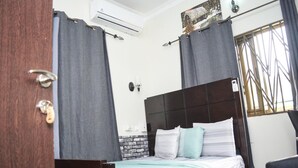 A bedroom with a bed, curtains, an air conditioning unit, a wall-mounted light, and a window with a metal grille. - 3A's Guest House (Akosombo)