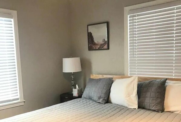 A king-size bed with a quilted, light-colored duvet in a room with neutral walls, a single bedside table with a lamp, and a framed picture on the wall. - Skots Villa Bottom West. Near Sanford Medical, Midco, Univ. SF & Augustana (Sioux Falls)