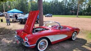 Un'elegante auto decappottabile rossa, con la capote abbassata, parcheggiata in un'area erbosa con alberi e altri veicoli sullo sfondo.