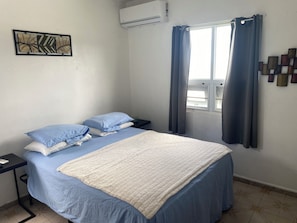 A bedroom with a bed, a nightstand, an air conditioning unit, a window with curtains, and a wall-mounted artwork.