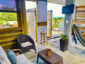 An open-plan living area with a sofa, a wicker chair, a small table, a stool, a TV, and a hammock.