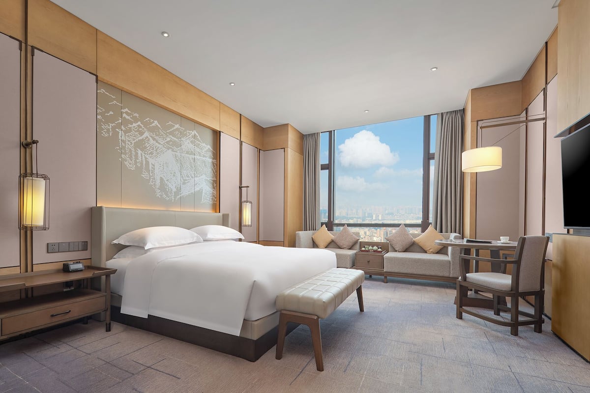 A modern hotel room with a large bed, a seating area with a table, and a city view through large windows.