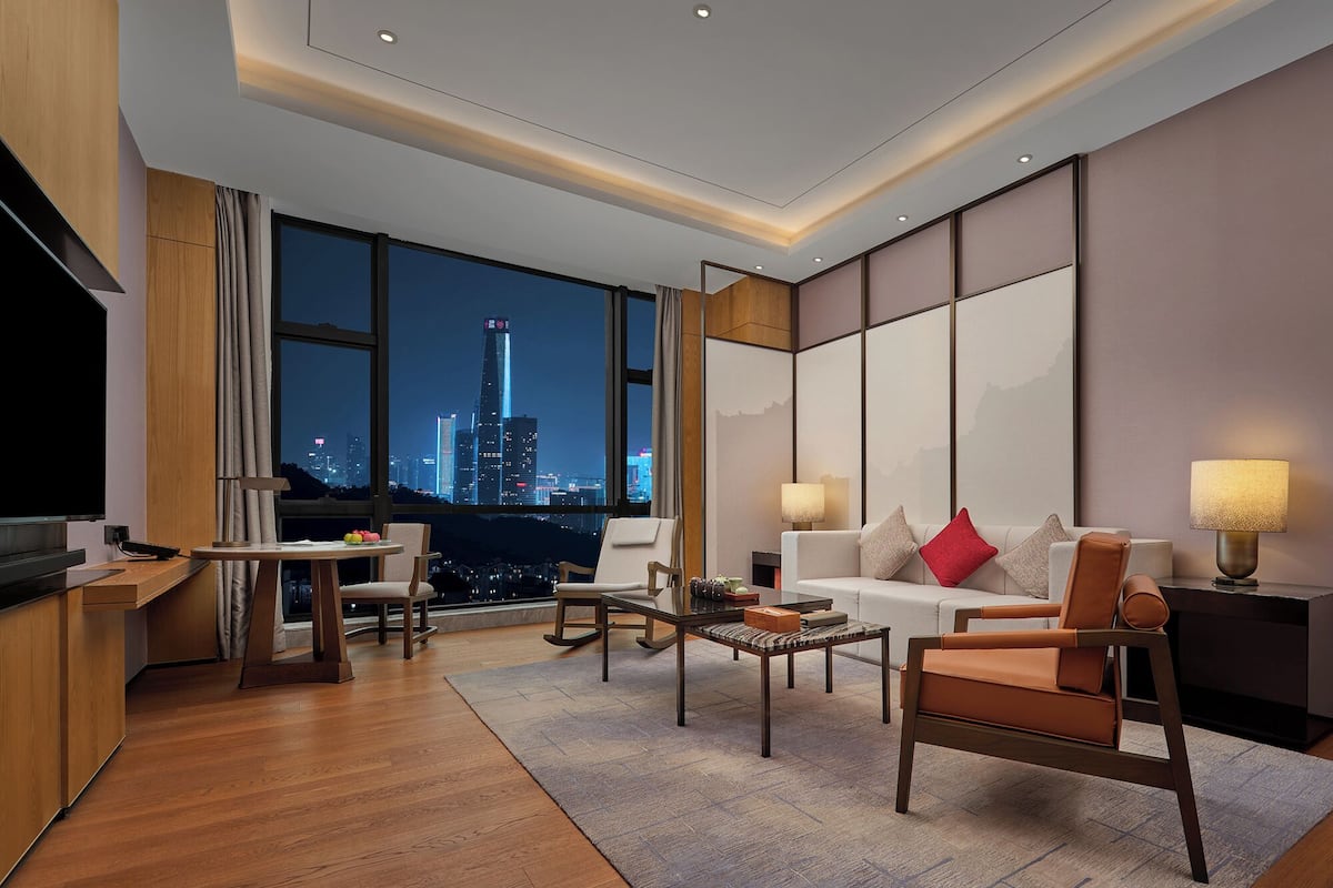 A modern hotel room with a city view, a flat-screen TV, a dining area, and a seating area with a sofa and chairs.