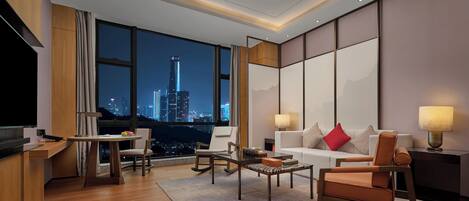 A modern hotel room with a city view, a flat-screen TV, a dining area, and a seating area with a sofa and chairs.
