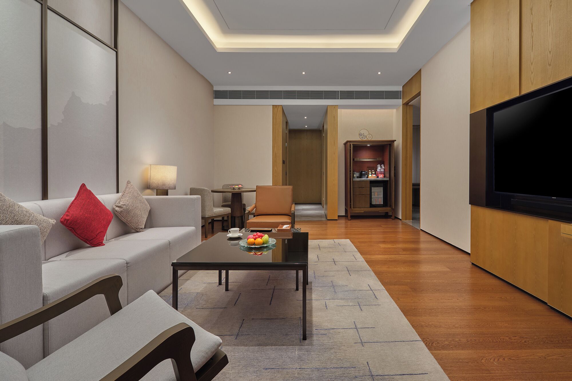 A modern living room with a sofa, coffee table, and TV. There is a dining area with chairs and a table. The room has wooden flooring and a large TV mounted on the wall.