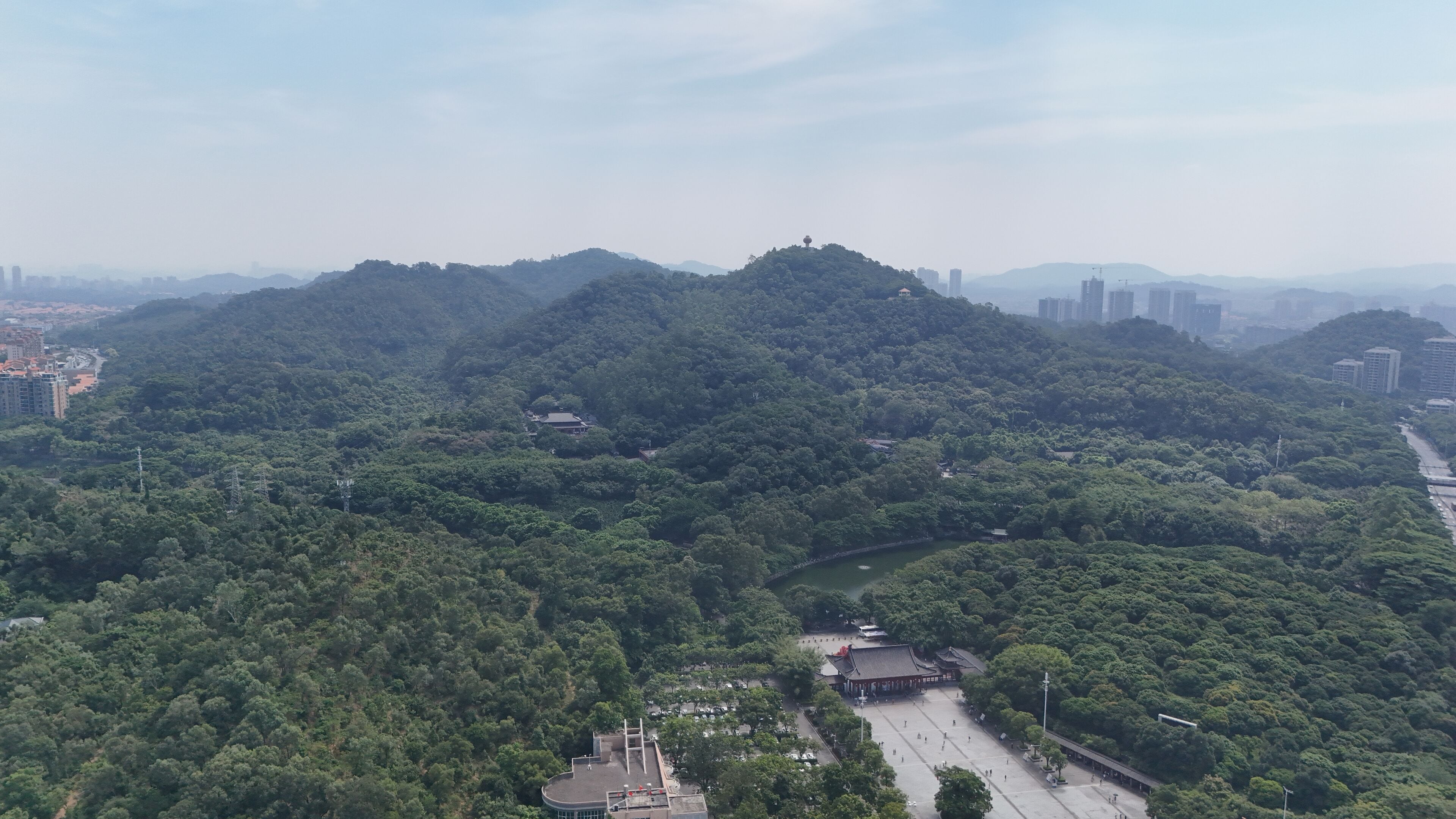 An aerial view of a lush green area with a traditional building complex, surrounded by dense forest and distant urban buildings.