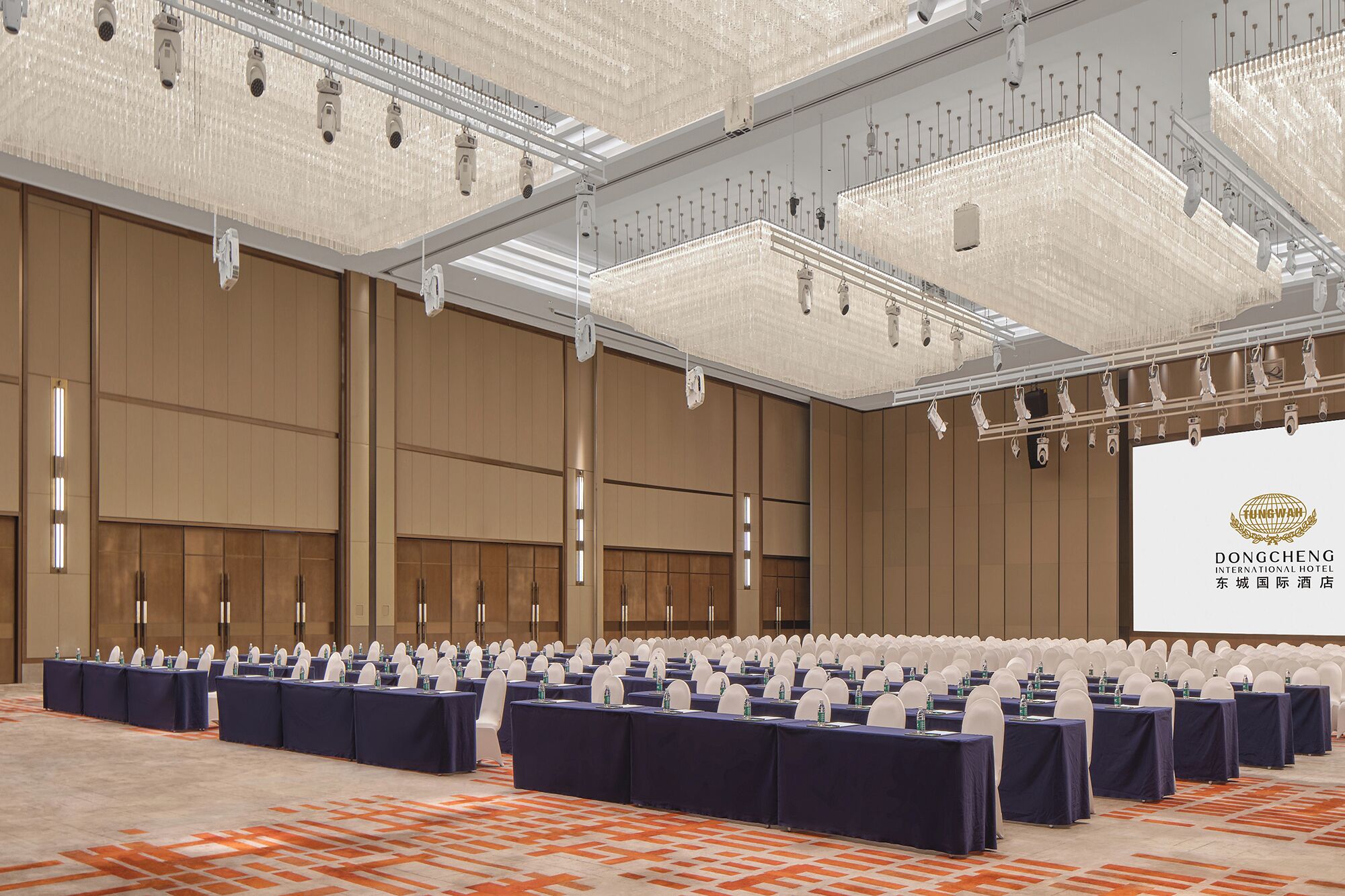 A large conference room with rows of tables and chairs, a large screen, and a chandelier.