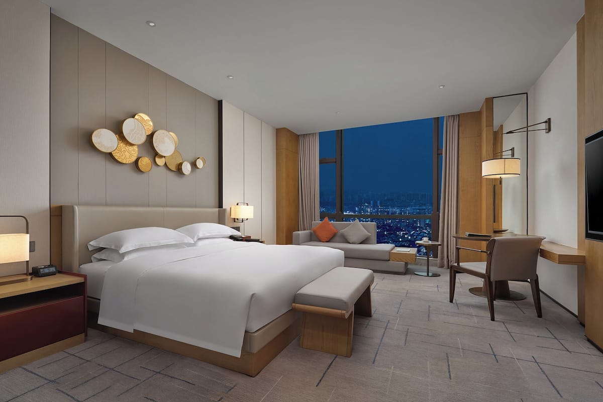 A modern hotel room with a large bed, a desk, and a view of the city at night.