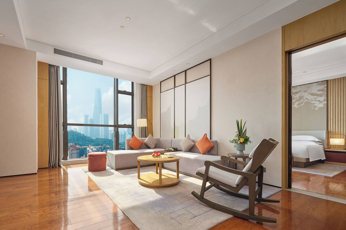 A modern hotel room with a large window offering a city view, a sofa set, a small round table, and a rocking chair.