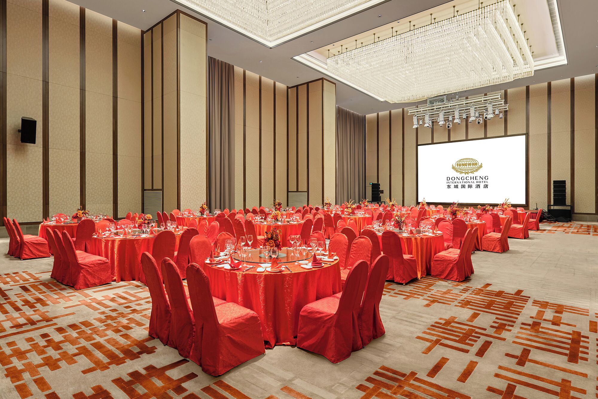 Un salón de banquetes con mesas redondas, sillas rojas y una pantalla grande que muestra el logotipo del Hotel Dongcheng International.