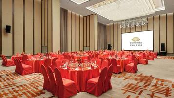 Un salón de banquetes con mesas redondas, sillas rojas y una pantalla grande que muestra el logotipo del Hotel Dongcheng International.