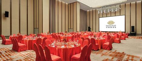Un salón de banquetes con mesas redondas, sillas rojas y una pantalla grande que muestra el logotipo del Hotel Dongcheng International.