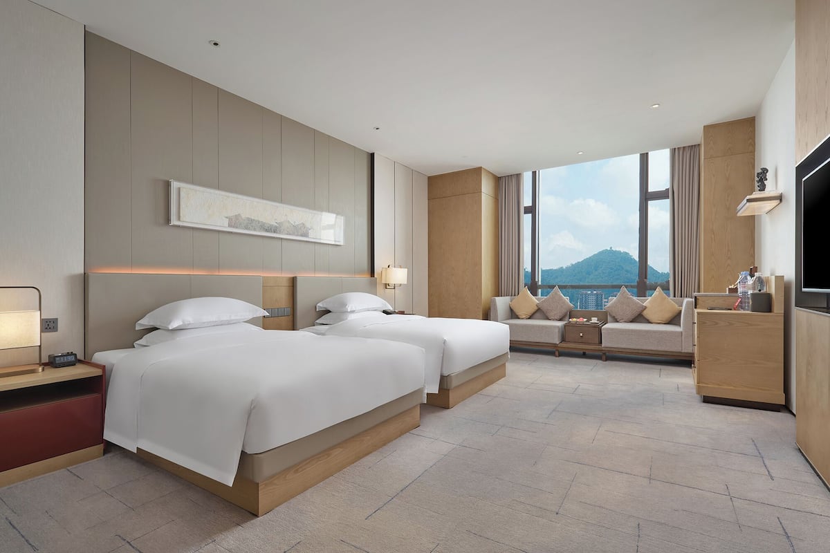 A hotel room with two beds, a sofa, a TV, and a large window with a view of a mountain.