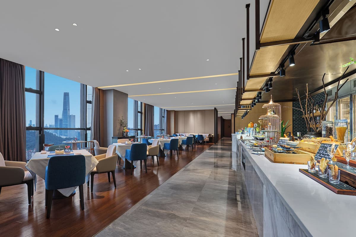A modern hotel dining area with long tables, chairs, and a bar counter with various food items.