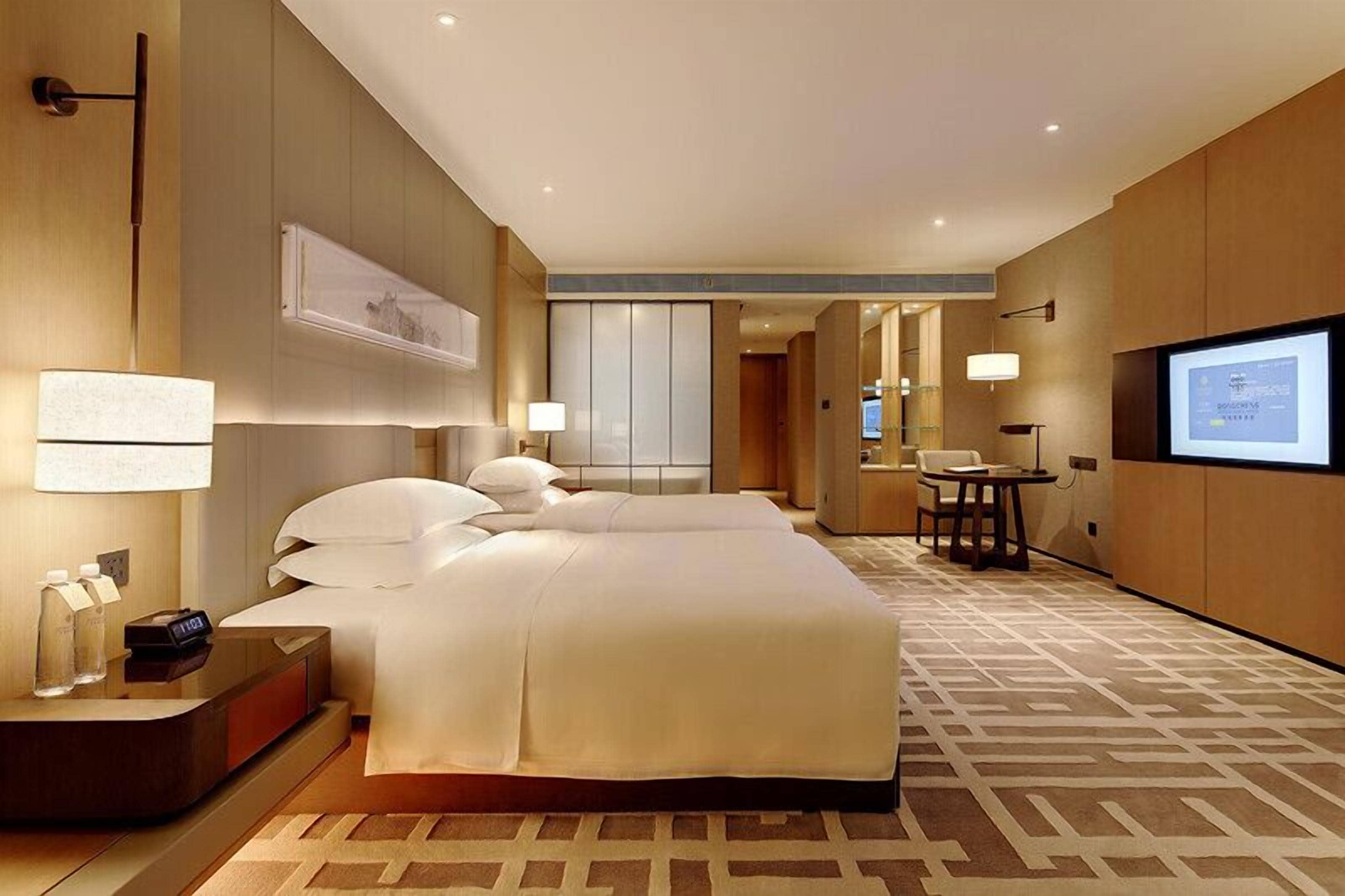 A modern hotel room with a large bed, bedside tables, a desk, and a TV screen.