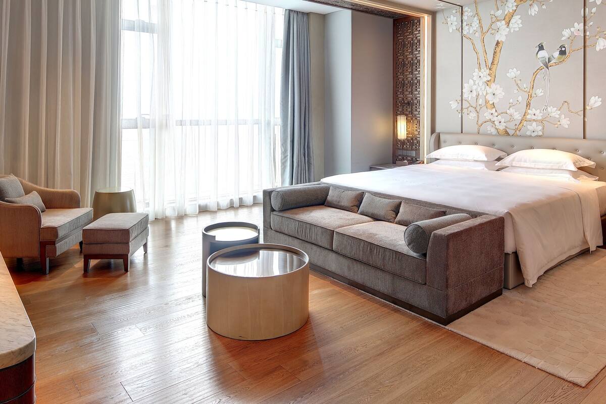Una habitación de hotel moderna con una cama grande, un sofá, una mesa redonda de centro y vistas al exterior a través de amplios ventanales.