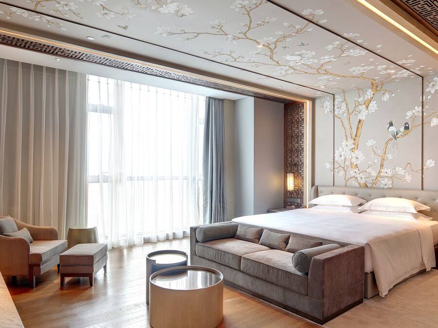 A modern hotel room with a large bed, a sofa, a round coffee table, and a view of the outdoors through large windows.